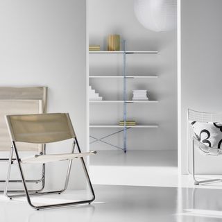 All white, sparse living room with an open door frame leading into another room. The front room has three accent chairs in neutral colors arrange around the doorway, and the back room has modern shelving against the wall and a paper lantern hanging from the ceiling.