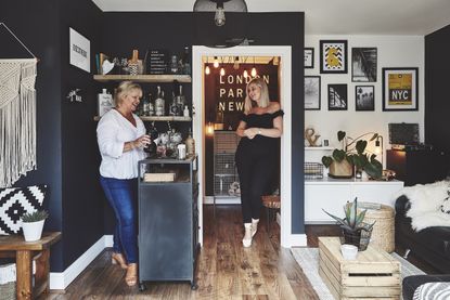 Mum and daughter stand at black freestanding bar cart with black painted wall, gallery prints and wooden flooring
