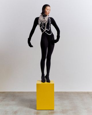 Woman standing on box in black bodysuit and pearl necklace