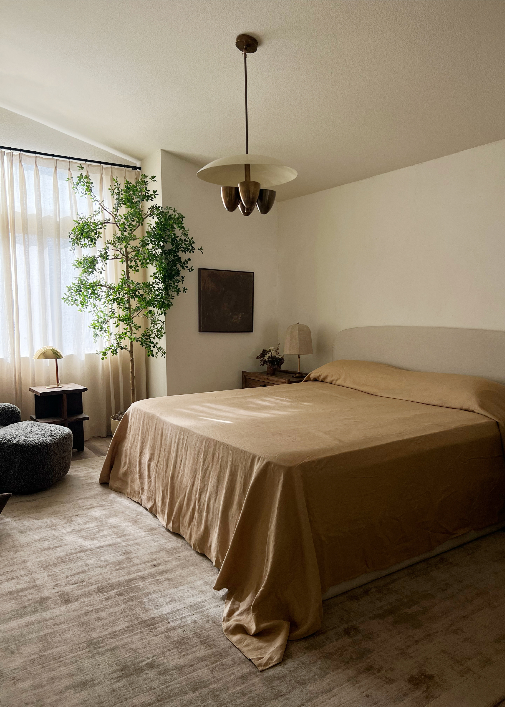 A beautiful bedroom with a minimalist bedscape, a statement pendant light, a potted tree, drawn sheer curtains, an armchair, and a side table with a table lamp