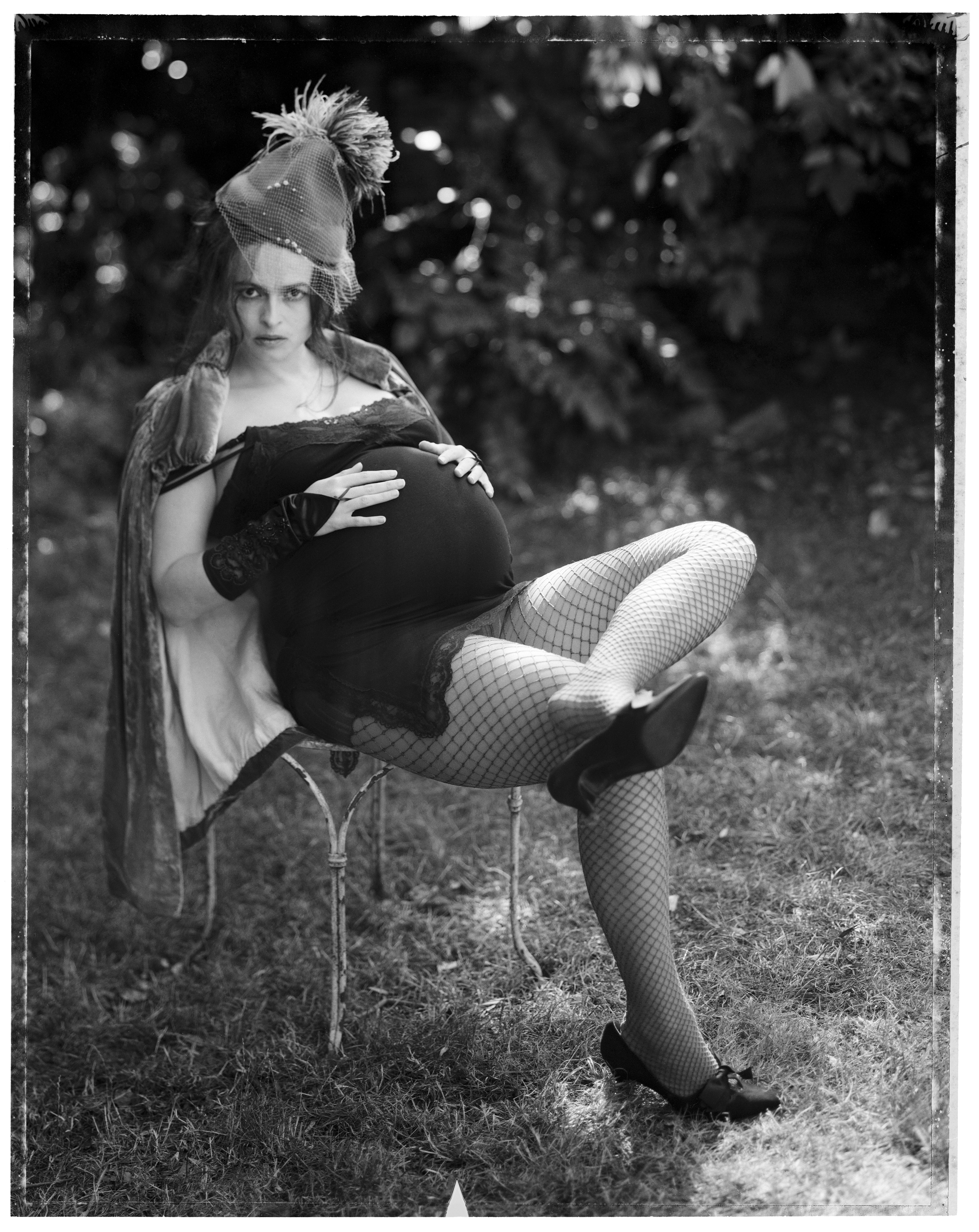 Helena Bonham Carter, visibly pregnant, sits on a garden chair in fishnet tights, heels and a feathered hat, one hand resting on her bump, looking directly into the camera.