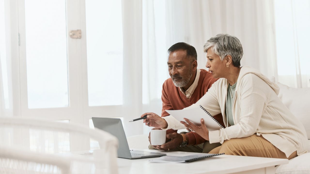 An older couple look at a laptop together while they go over paperwork on their living room sofa.