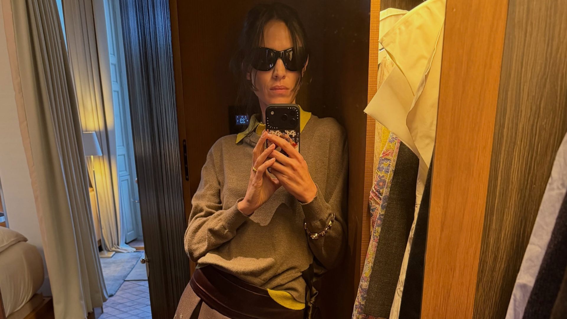 Alexa Chung takes a mirror selfie wearing a knee-length pleated skirt with a grey jumper, a yellow shirt and a purple cummerbund.