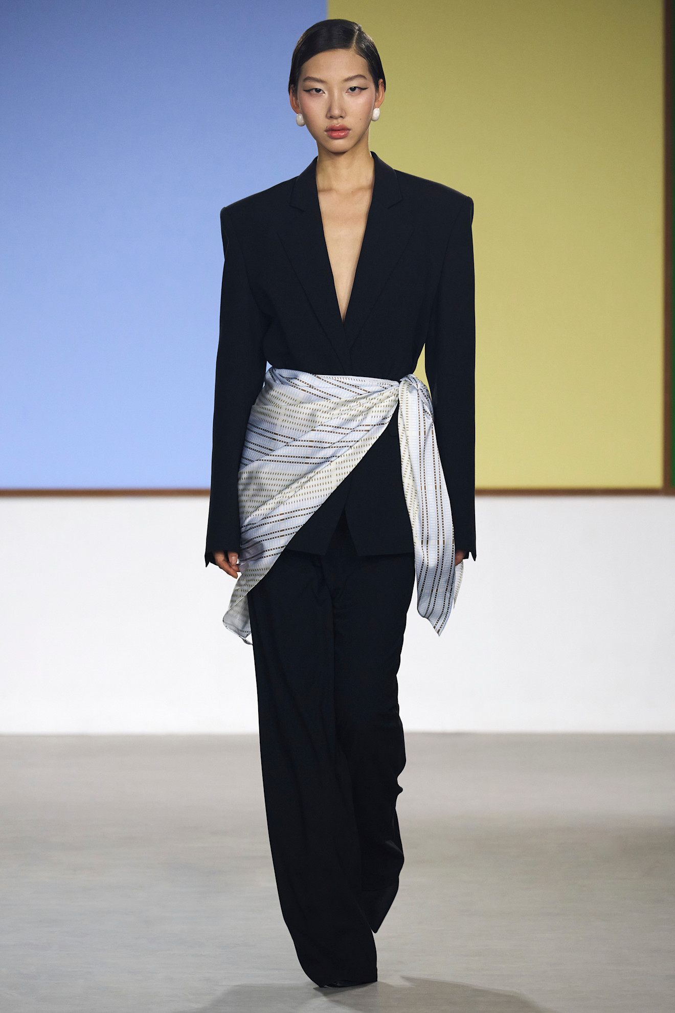 Layering staples for spring 2026 is shown in a photo from Yirantian's runway show, with a model walking while wearing a black blazer styled with black trousers, heels, silver earrings, and a light blue printed silk scarf tied around the waist