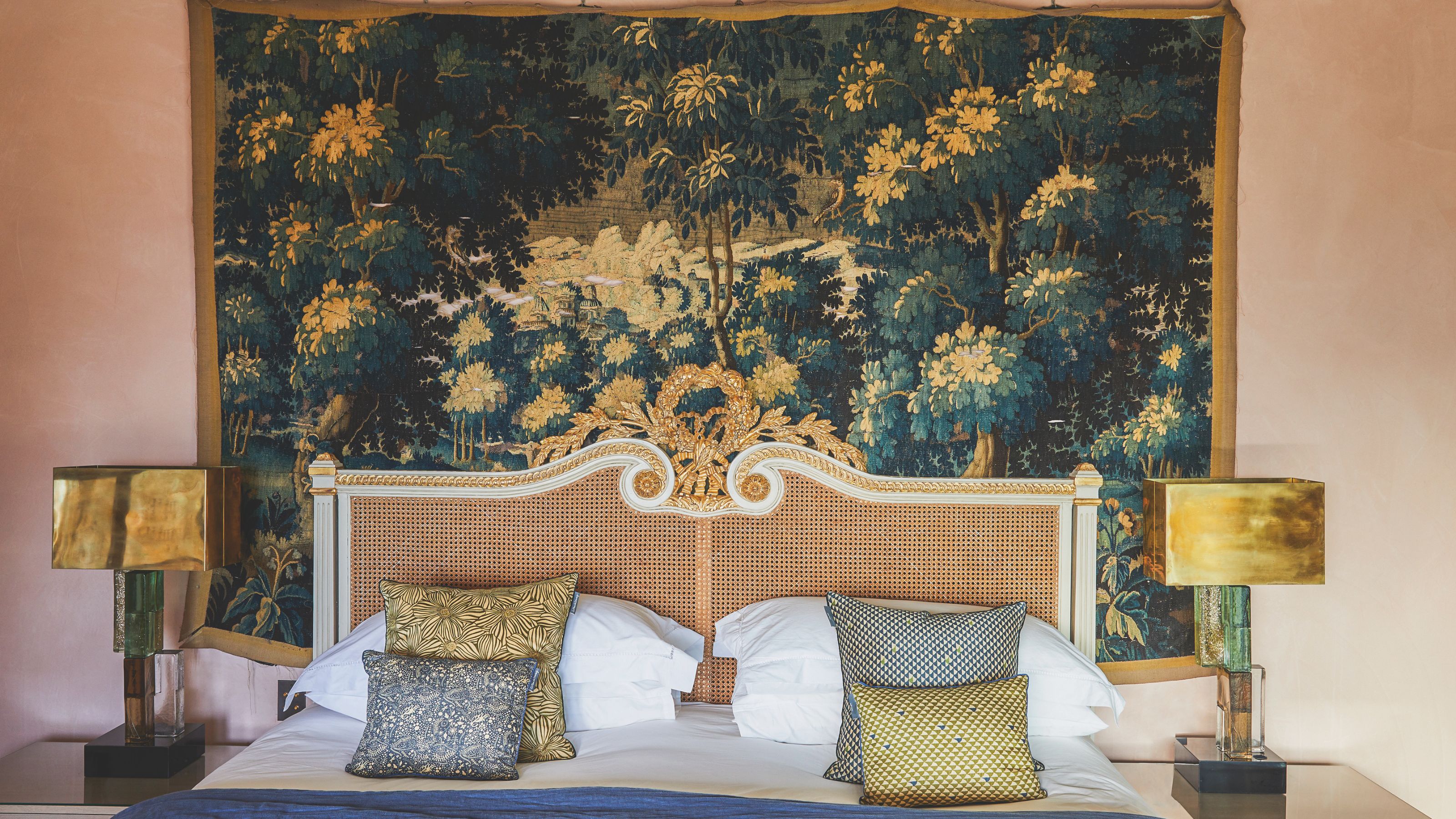 Ornate cane bed with gold trim against a large forest tapestry backdrop, flanked by brass lamps