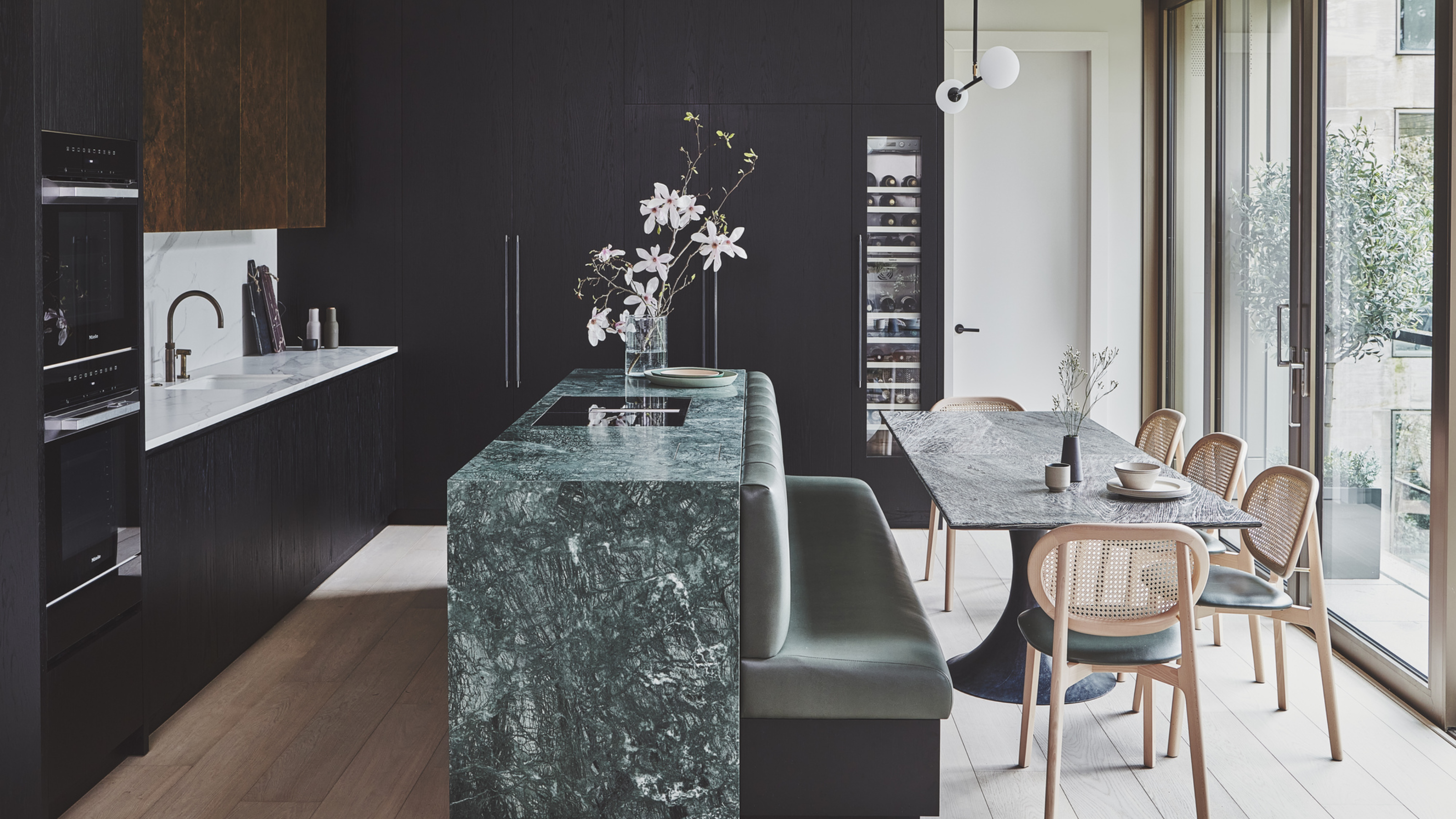 A kitchen with black cabinetry, an island with built-in banquette seating, a dining table, and a wine fridge with a glass door built-into the cabinets