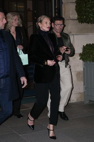 Kate Moss wears a sheer top, black blazer, black pants, and black front strap mule pumps.