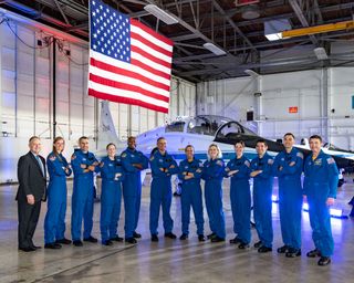 a bunch of people standing in blue flight suits in front of a t-38 jet and an american flag, in a hanger