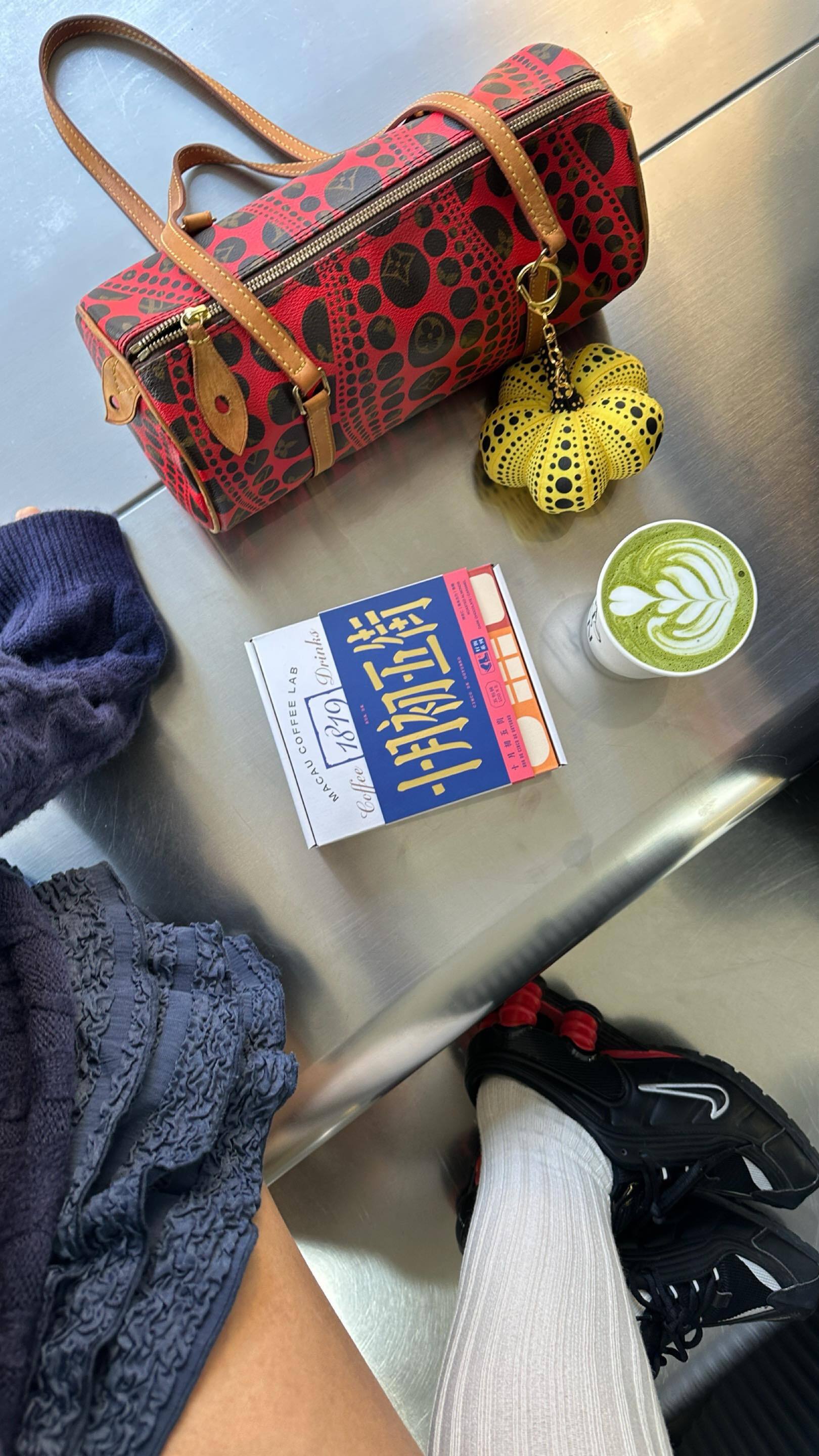A Louis Vuitton bag and matcha latte on a bench next to a woman wearing long white socks and black Nike Shox sneakers.
