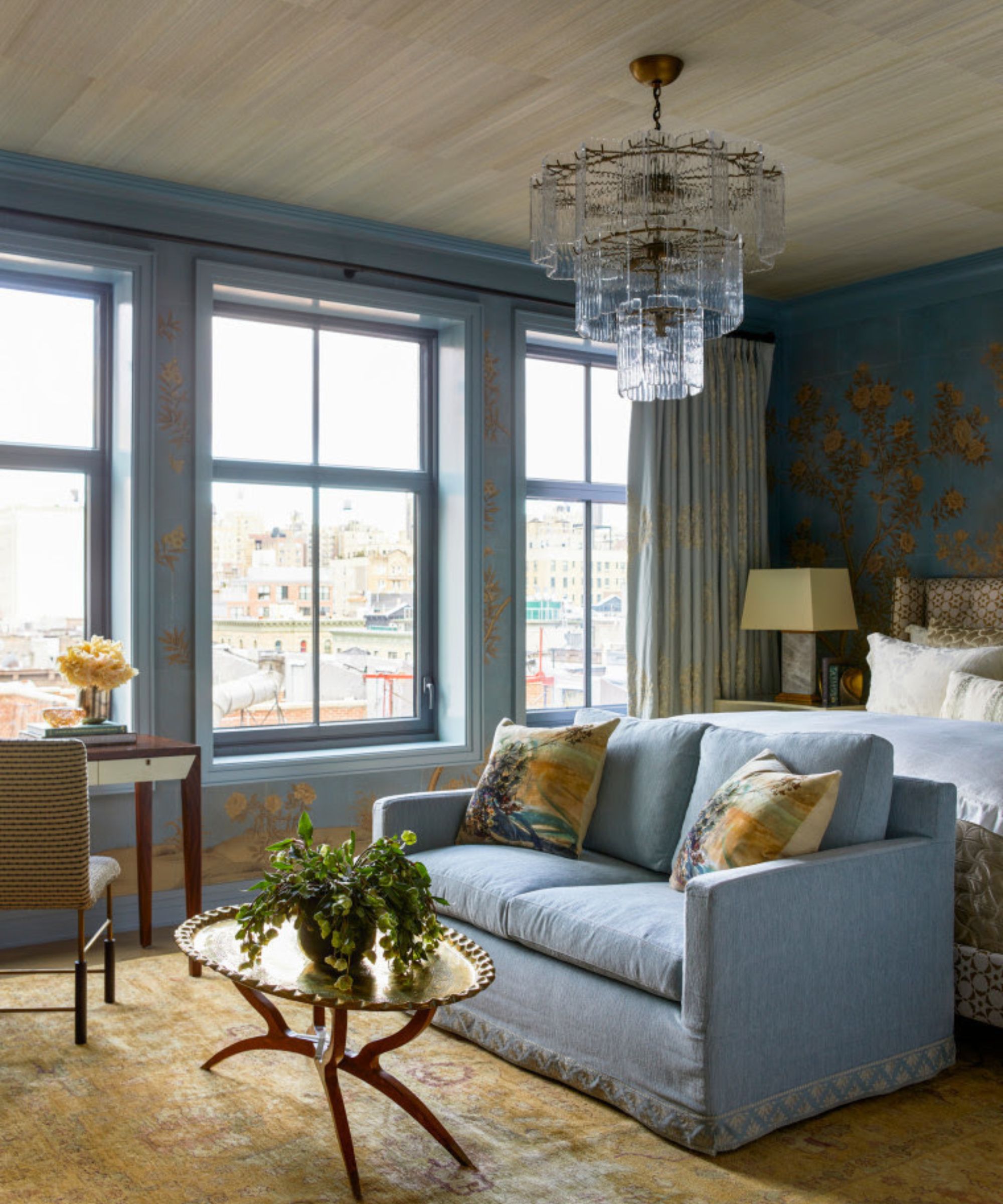 A luxurious blue bedroom featuring a light blue loveseat, a gold tray coffee table, and blue floral wallpaper. A tiered glass chandelier hangs from a textured ceiling, and large windows offer a city view.