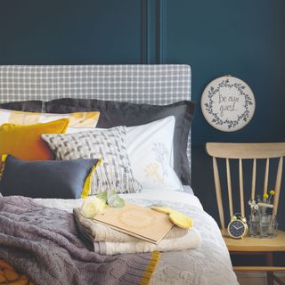 Blue panelled guest bedroom with lots of cushions and blankets on the bed and a wooden chair with a clock and flowers on it at the side