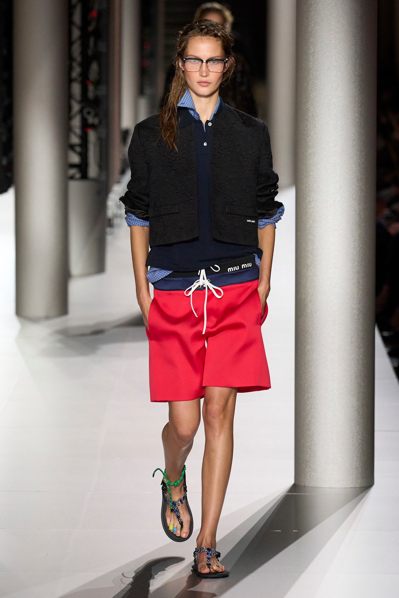 a Miu Miu Spring 2024 model wearing red board shorts, a blue button down, a blue polo shirt, a black blazer, and rope sandals on the runway