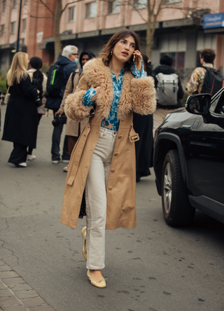 Jeanne Damas talks on the phone and wears a tan leather jacket with a shearling collar, blue print shirt, white pants, and light yellow ballet flats at the Coperni show during Paris Fashion Week Fall/Winter 2022 on March 03, 2022 in Paris, France