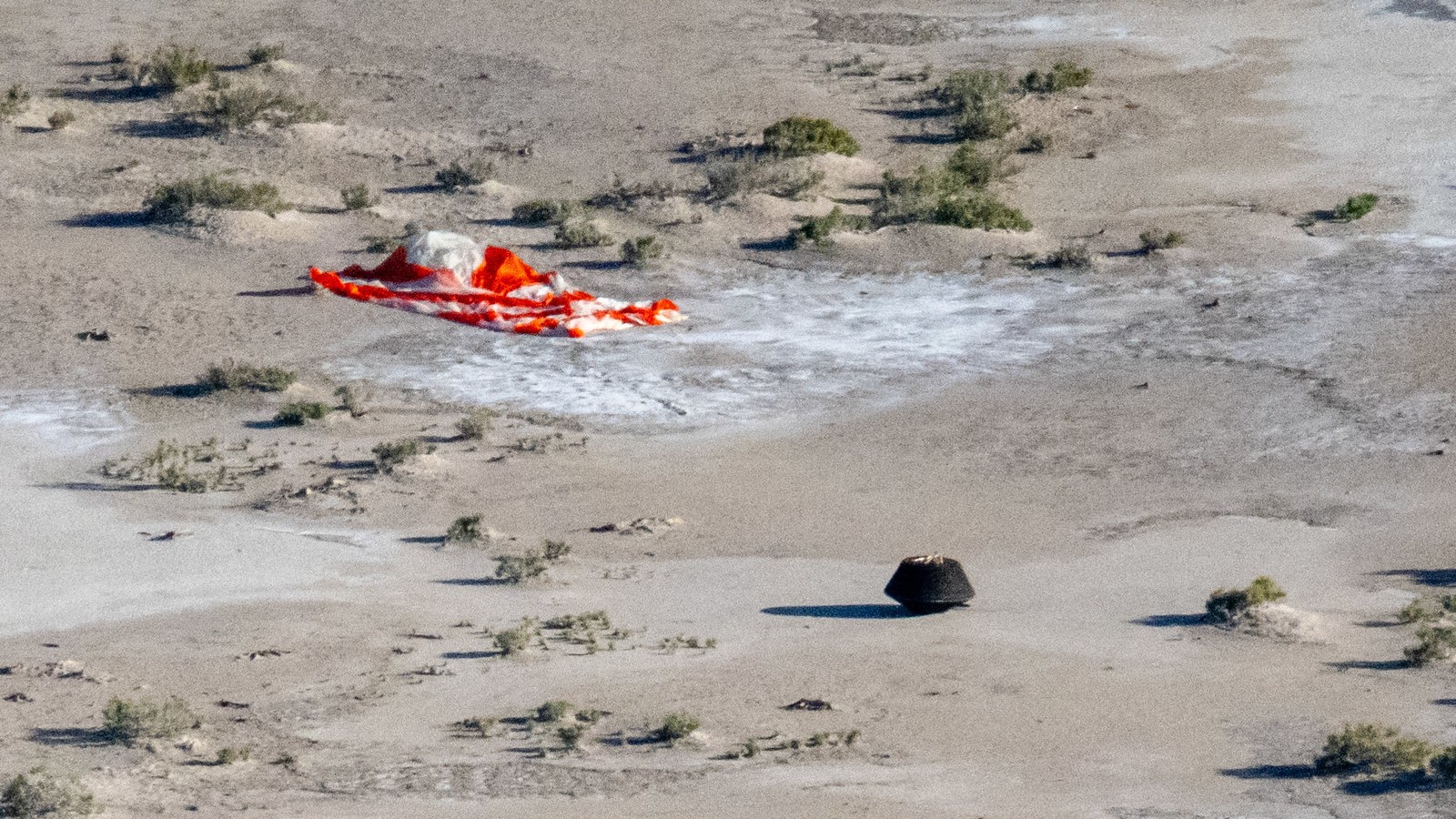 a black cone-shaped capsule sits in the desert beside an orange parachute