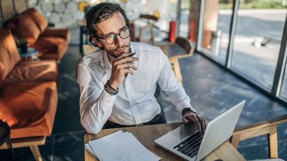 A business owner looks thoughtful while sitting at a table and using a laptop.