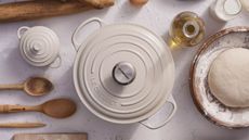 A cream colored Le Creuset Dutch oven viewed from above, with a small cocotte version beside it. With wooden baking utensils, and a bowl of bread dough beside it. 