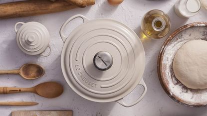 A cream colored Le Creuset Dutch oven viewed from above, with a small cocotte version beside it. With wooden baking utensils, and a bowl of bread dough beside it. 
