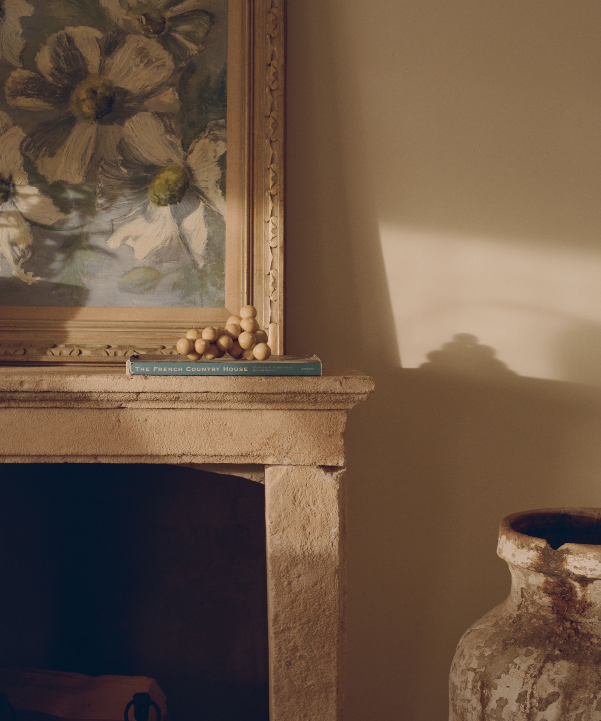 Close-up of a stone fireplace mantle holding a book, marble grapes, and a framed floral painting in warm sunlight.