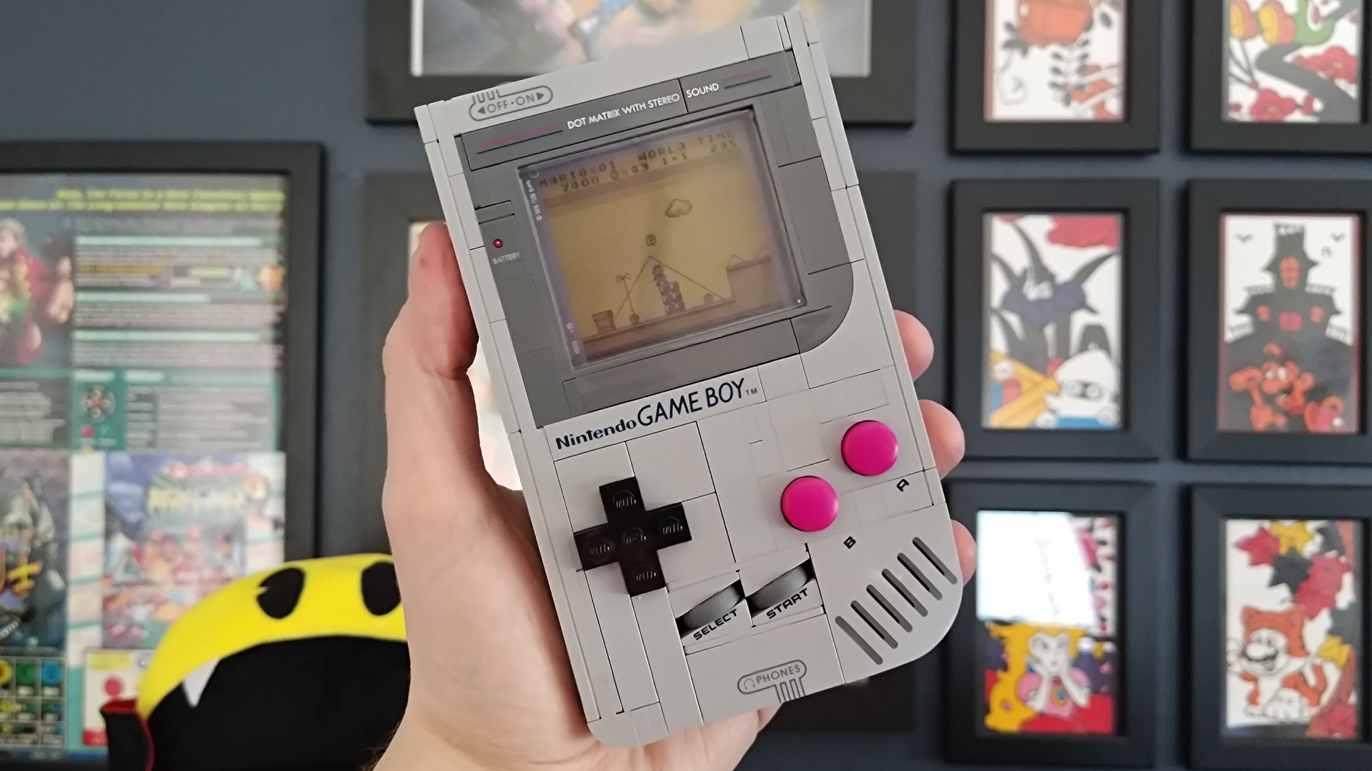 A hand holds up the Lego Game Boy, with retro posters visible in the background