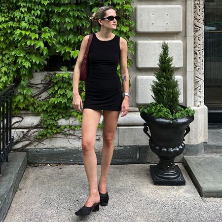 Eliza Huber wearing a black Norma Kamali mini dress with black mules, a burgundy bag, sunglasses, and a slicked back bun.