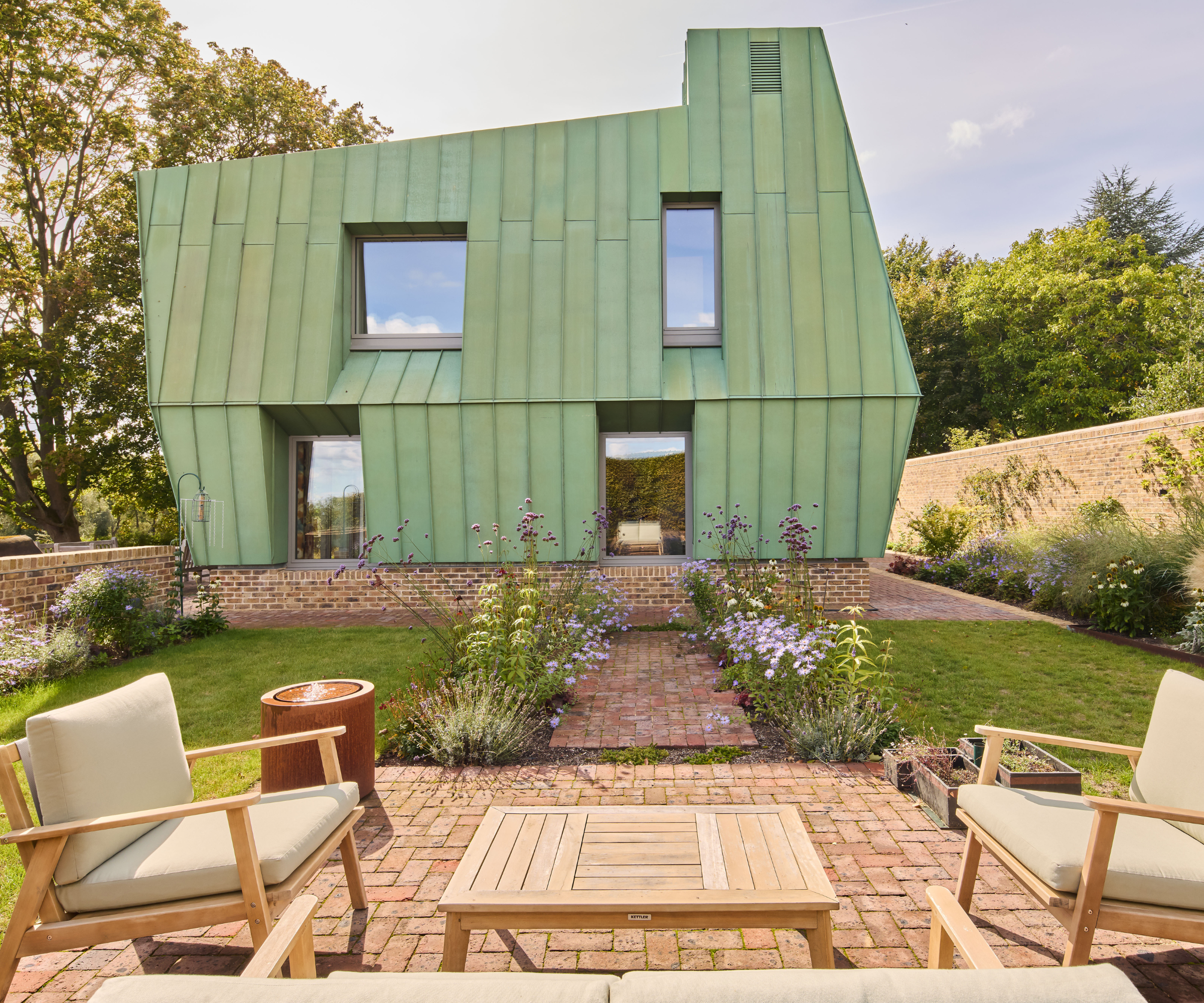 Garden area in front of green house with brick patio and seating with a walkway with flowers on either side