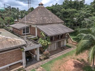 Daytime, exterior image of Nwoko’s home-cum-studio in Idumuje-Ugboko shot from the air, brick building, brick pointed roof, windows, doorway and porch area, steps, motorbike parked, tall green trees, flowers, stone flags, scorched lawn, blue cloudy sky