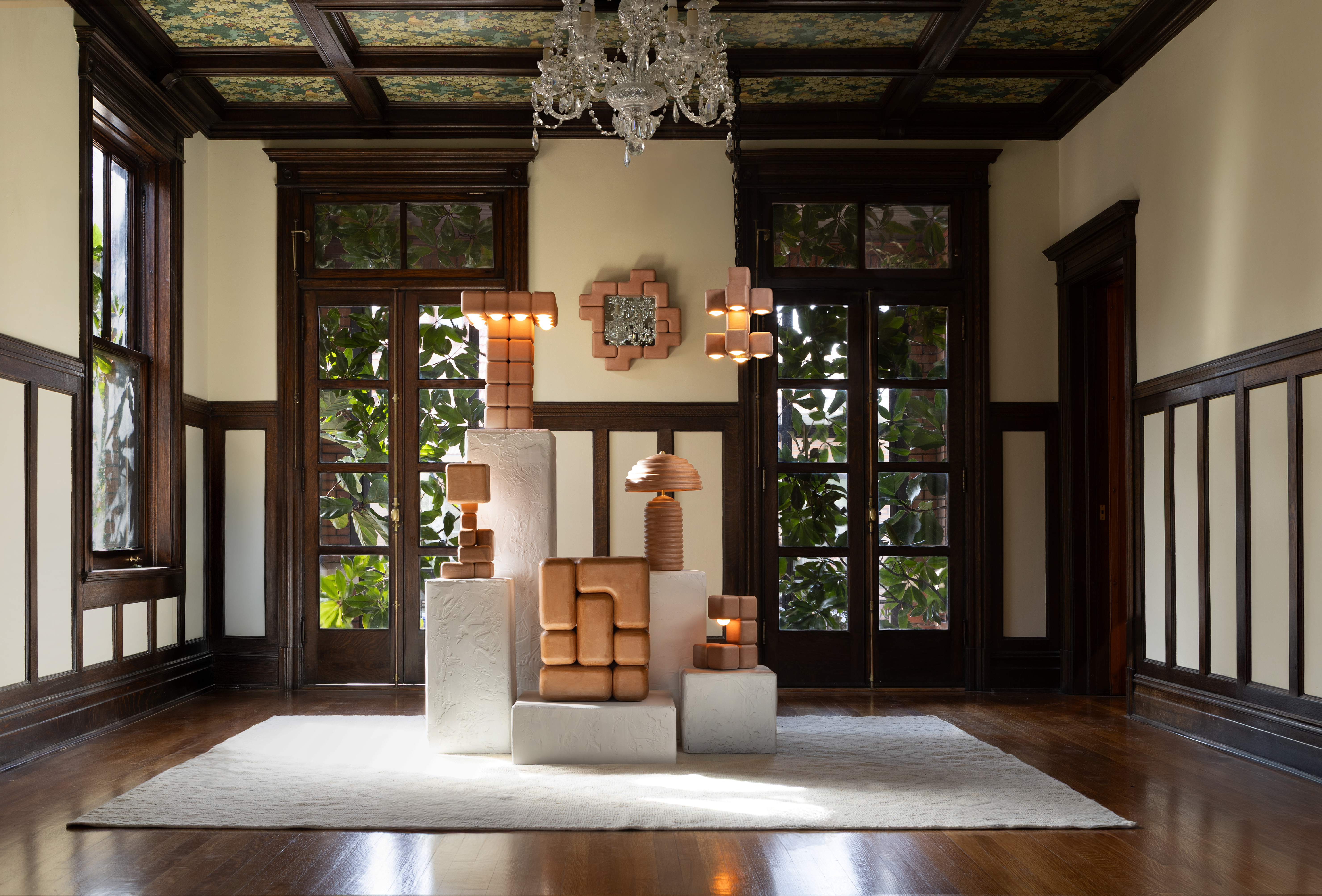 A series of whimsical, soft-shaped ceramic lamps and sculptures sits atop white podiums positioned, in turn, over a white rug in a wood-drenched room with buttery walls.