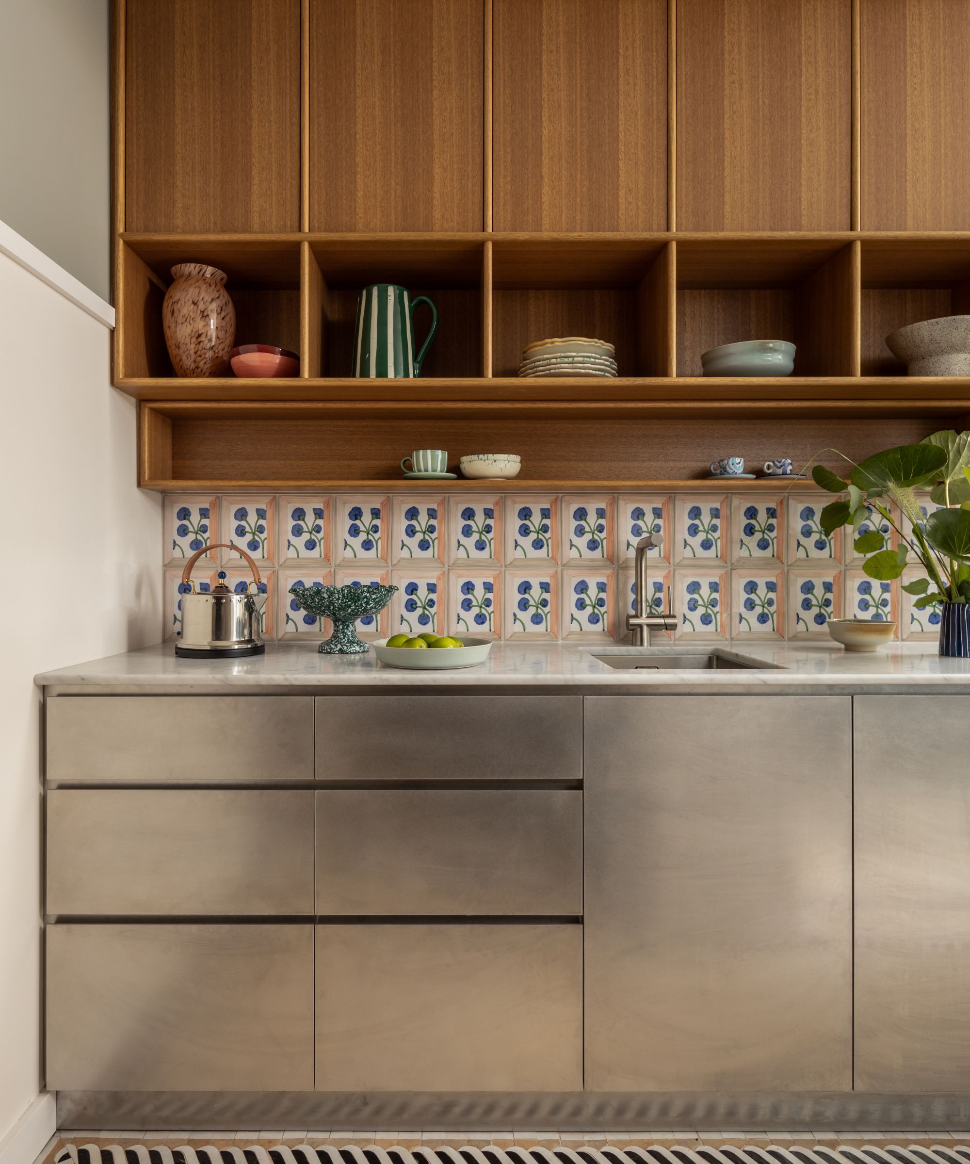 A modern kitchen counter featuring brushed stainless steel cabinets and a white marble countertop. The backsplash consists of decorative hand-painted floral tiles, set beneath warm wood open shelving filled with ceramic bowls and glassware.