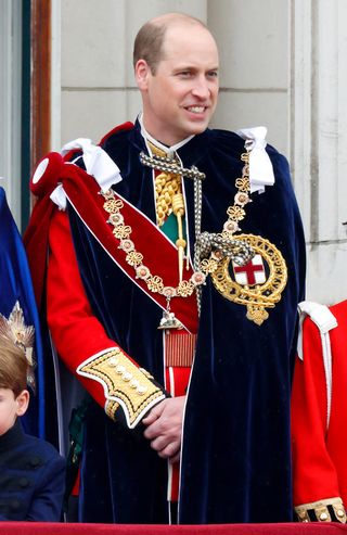 Prince William wearing a red uniform and coronation robe with medals smiling with his hands clasped in front of him