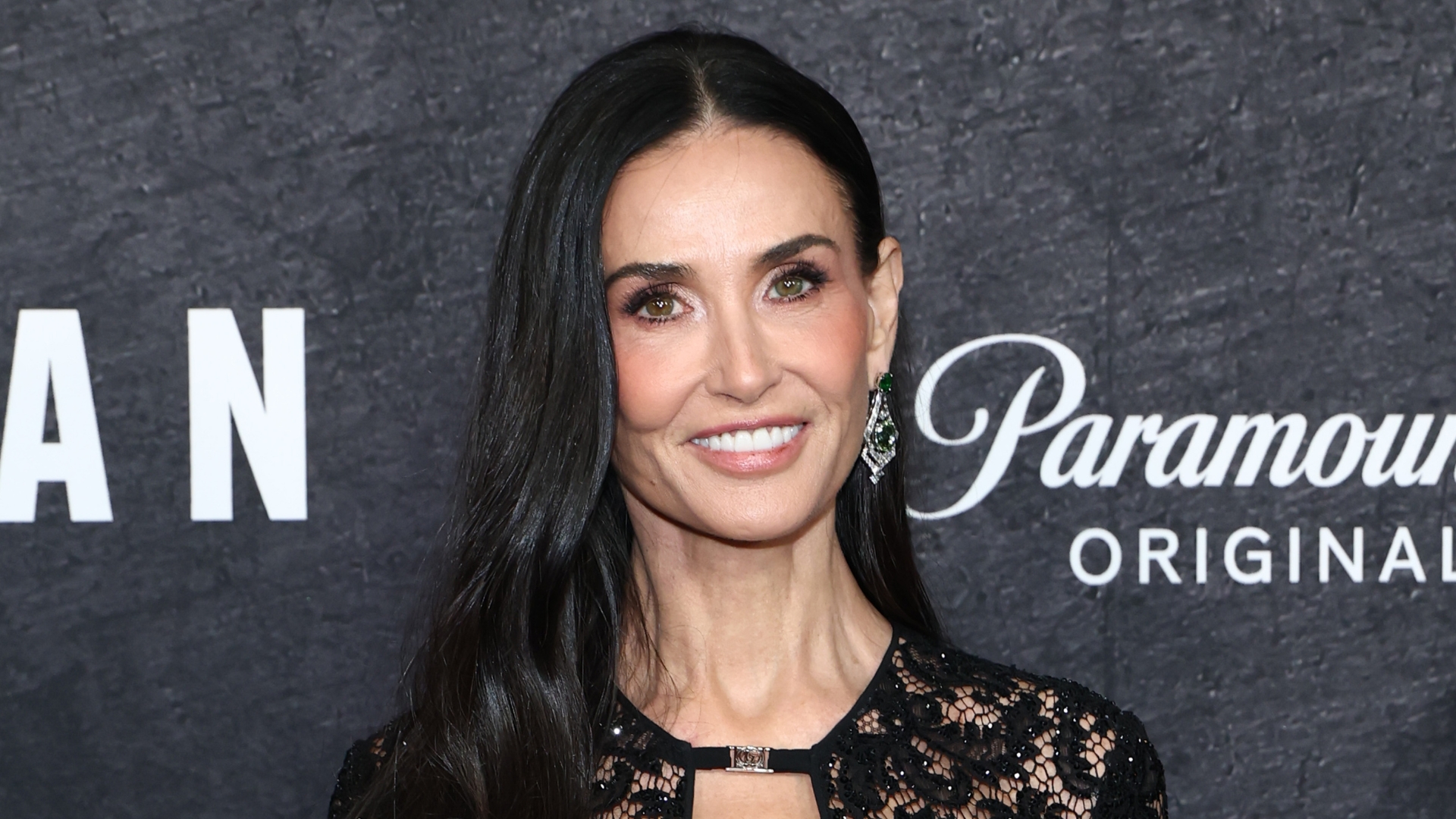 Image of Demi Moore smiling with long black hair curled over one shoulder, and wearing a black lace gown