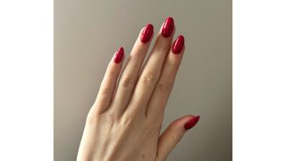 A close-up of Digital Beauty Writer, Sennen Prickett's hand, which features a cranberry-red, almond shaped manicure - pictured in front of a cream background