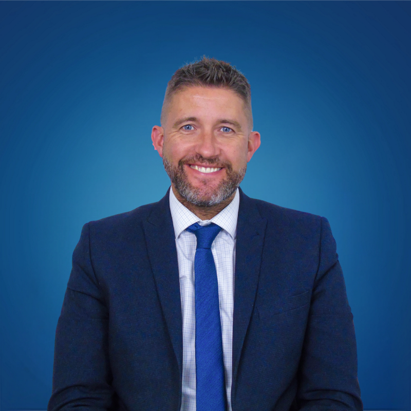 male with short dark grey hair and beard wearing blue tie, white shirt and navy suit sat against blue background
