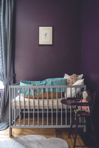 baby girl nursery with eggplant colored walls, moses basket, chair with doll, gray drapes