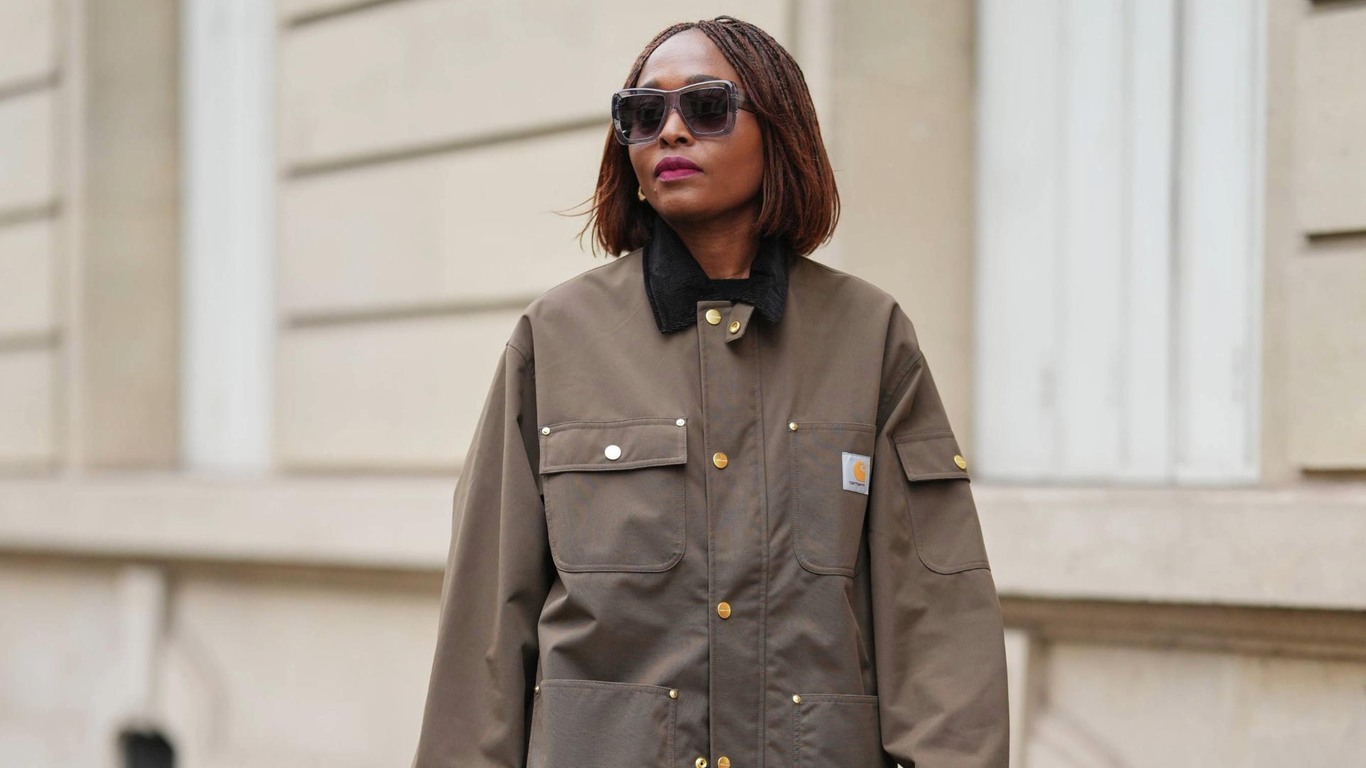 PARIS, FRANCE - DECEMBER 13: Carrole Sagba wears a straight chin-length bob hairstyle with a center part, makeup featuring a natural complexion with soft pink lipstick, an olive green khaki cotton canvas workwear jacket with flap chest pockets and snap-button fastening by Carhartt, black opaque tights, a burgundy leather tote bag with a structured silhouette and crocodile pattern, and burgundy shiny knee-high leather boots with a pointed toe and block heel and crocodile pattern, during a street style fashion photo session, on December 13, 2025 in Paris, France. (Photo by Edward Berthelot/Getty Images)