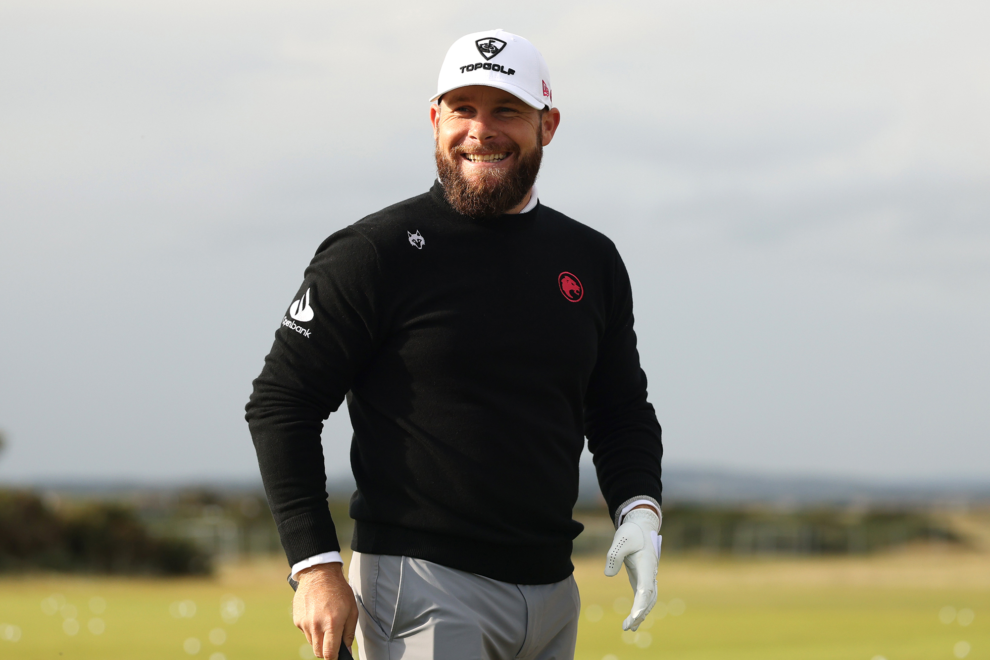 Tyrrell Hatton smiling on the golf course, holding a golf club in his right hand