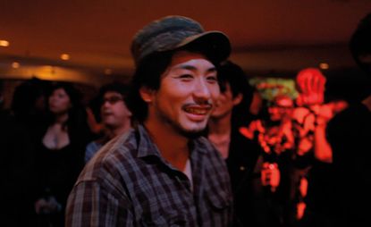 View of a man dressed in a dark coloured plaid shirt and a hat at a venue. There are multiple people in the background