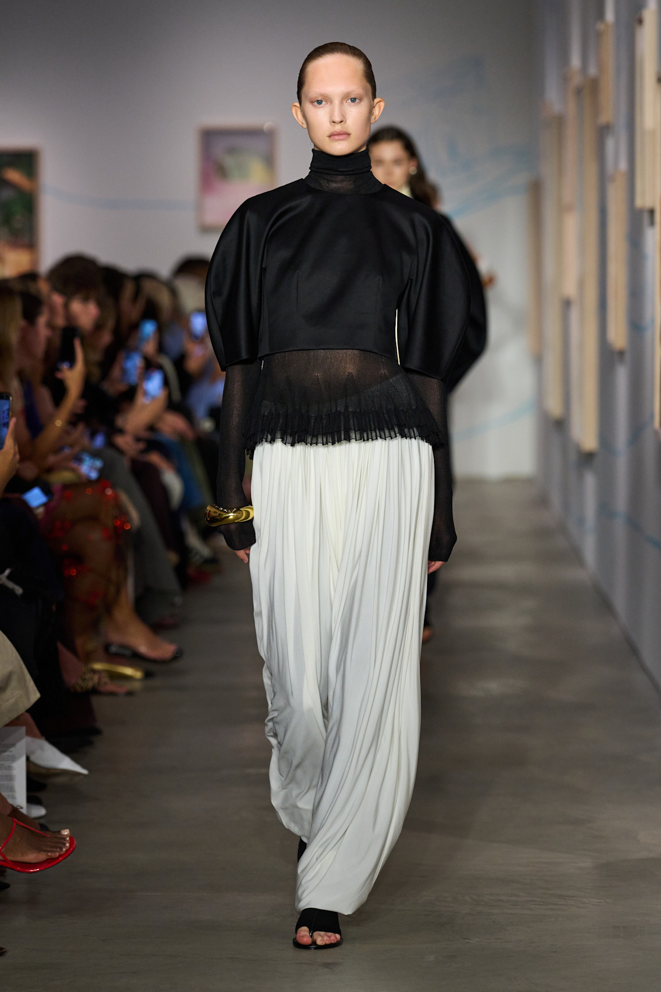 Layering staples for spring 2026 is shown in a photo from Ashlyn's runway show, with a model walking while wearing a black satin cropped puff sleeve blouse over a sheer black turtleneck shirt styled with white pleated pants, black flat sandals, and a single gold bangle