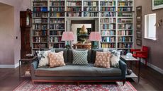 Living room with built-in bookcase, grey sofa, red Persian rug, grandfather clock, two lamps with pink shades and lime wash walls