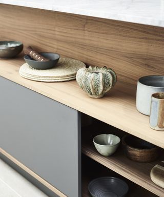 A close up of wooden open shelving in an island with grey panelling, and a collection of small bowls in natural materials
