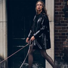 model wears patent barn style jacket, a skirt, and tights while walking a dog