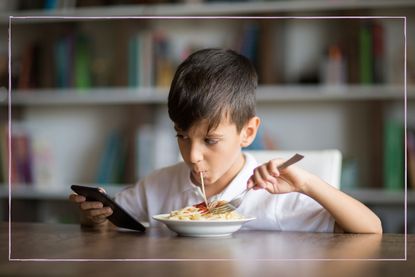 A young boy on his phone while eating spaghetti