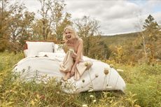 Naomi Watts on a bed with beige bedding in the middle of a wild, grassy field