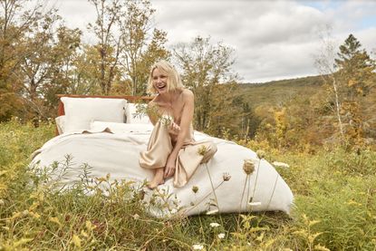 Naomi Watts on a bed with beige bedding in the middle of a wild, grassy field