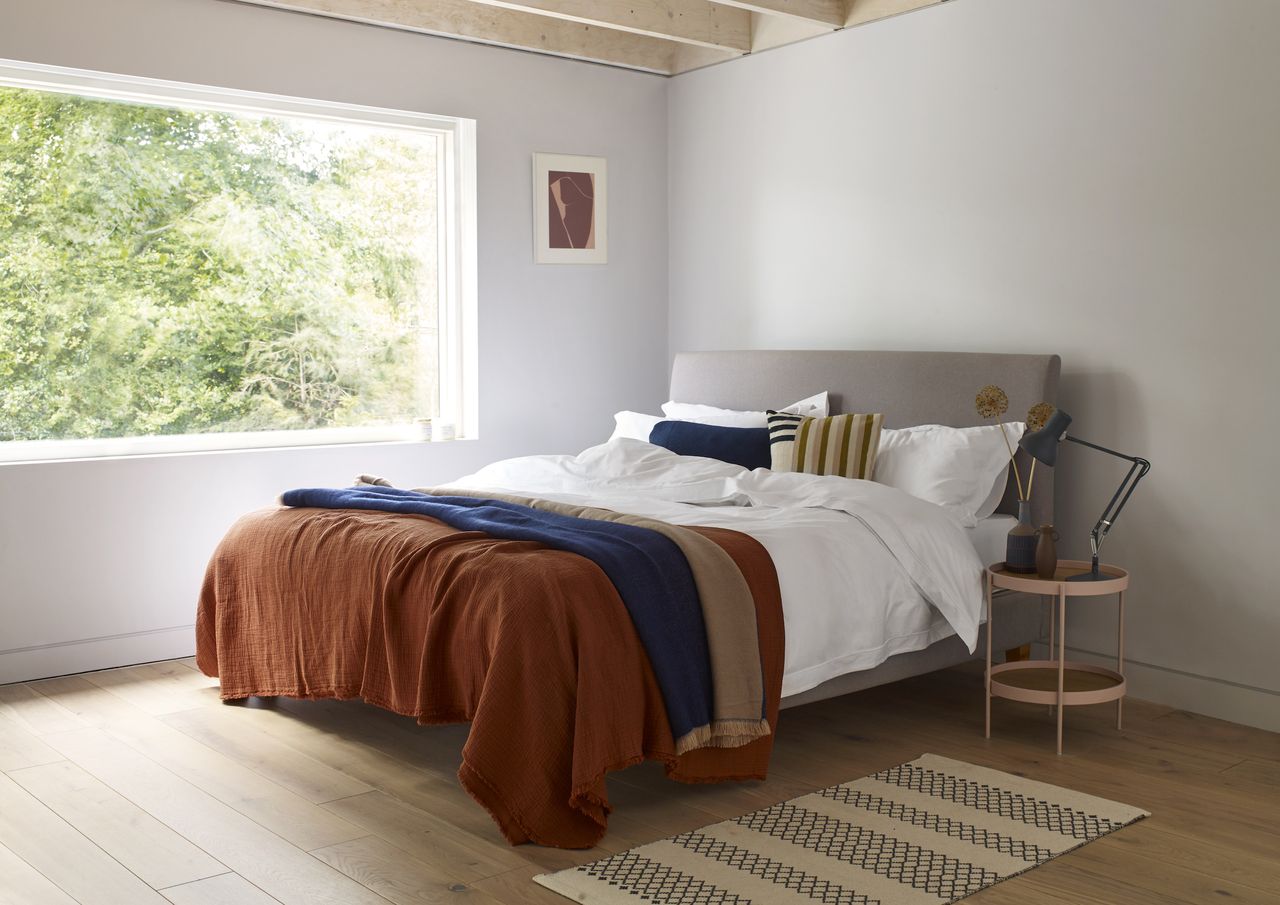 bedroom with gray double bed, hardwood floor, pink side table with blue lamp, rust and blue blankets and cushions, large window, artwork, rug