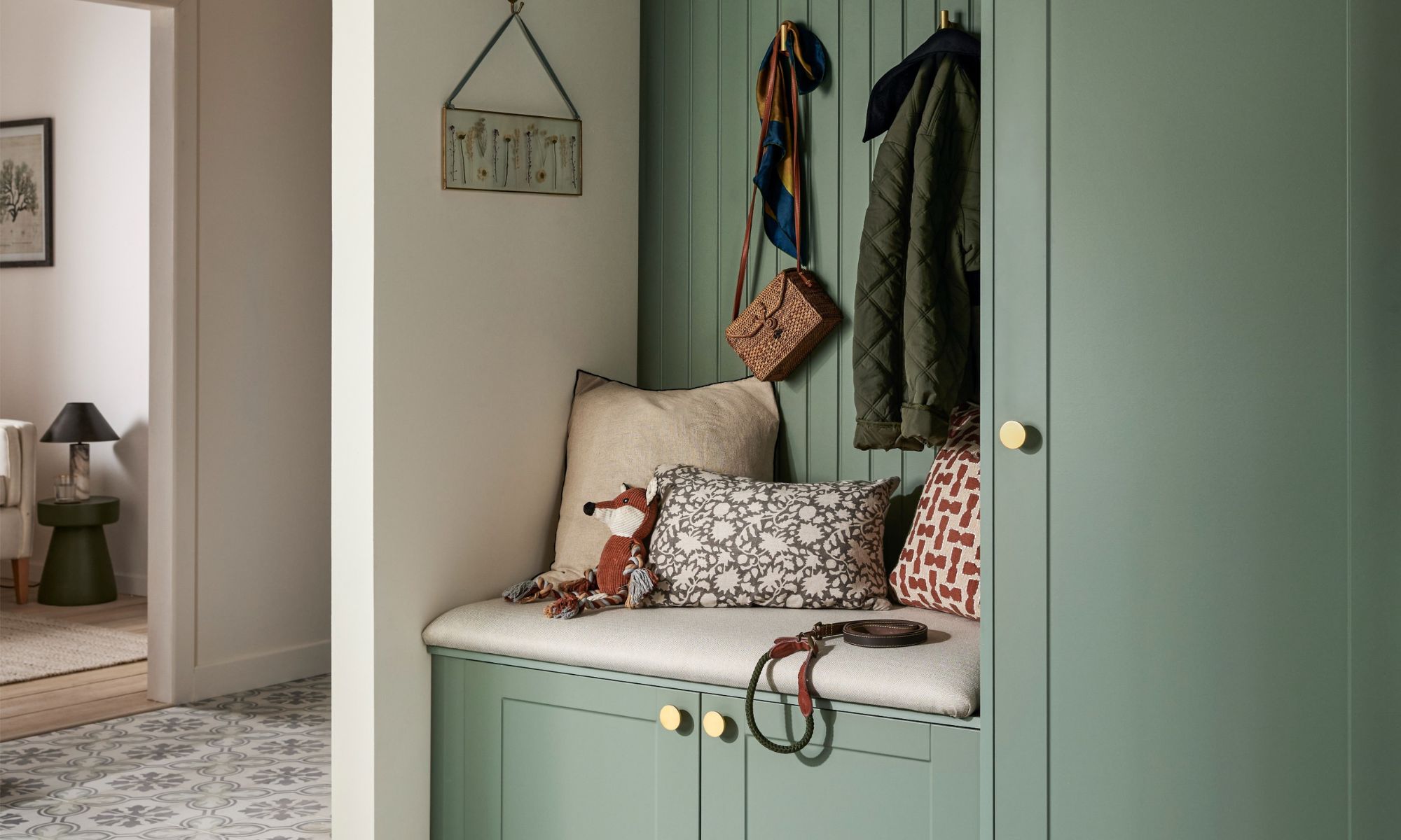 A sage green boot room with a cushioned storage bench, brass knobs, tongue-and-groove panelling, brass coat hooks holding a quilted jacket and wicker bag, and a dog lead