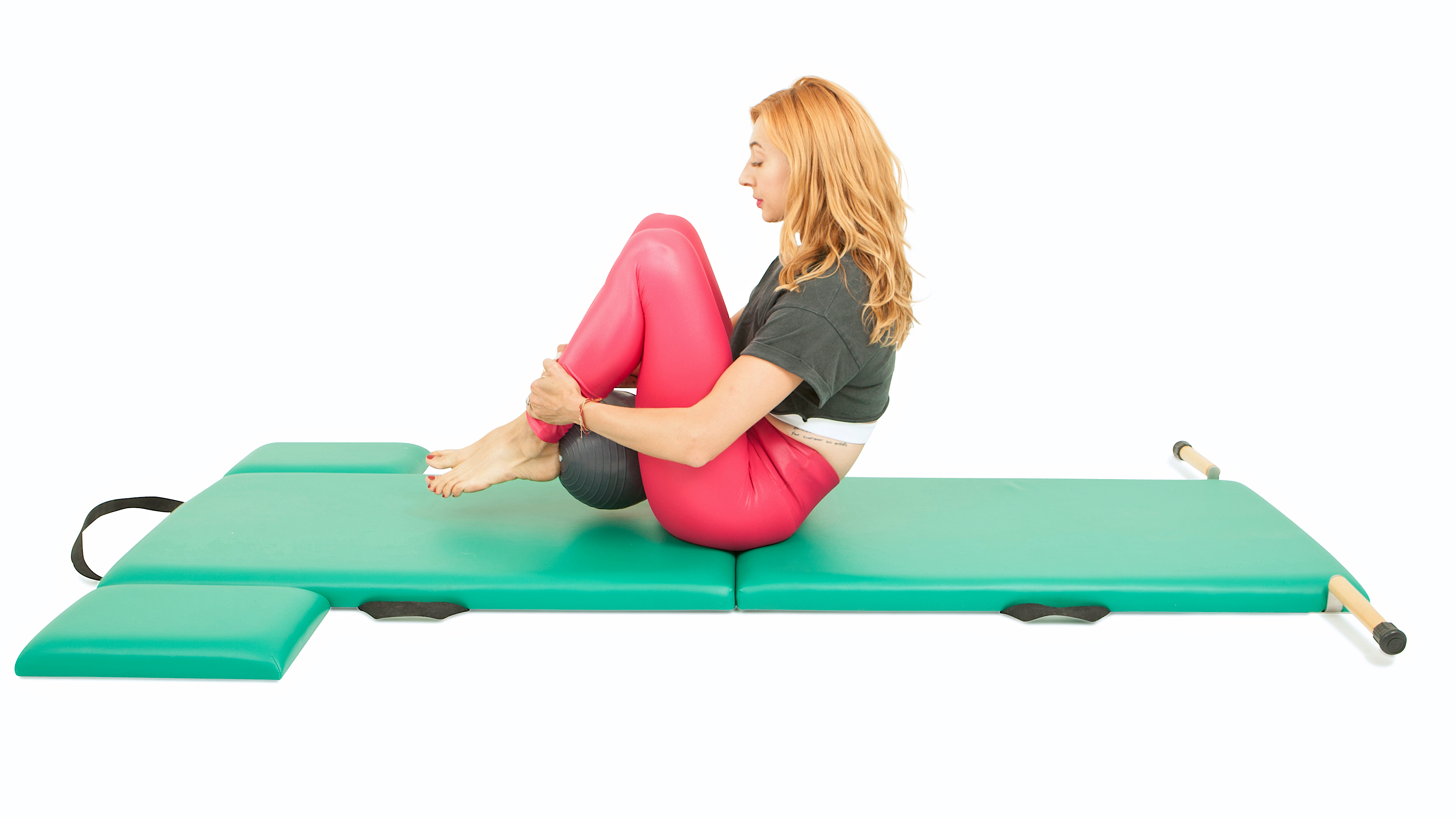Lesley Logan sits on a padded mat, with her knees bent and feet hovering off the floor. A Pilates ball is held between the heels of her feet and her buttocks.
