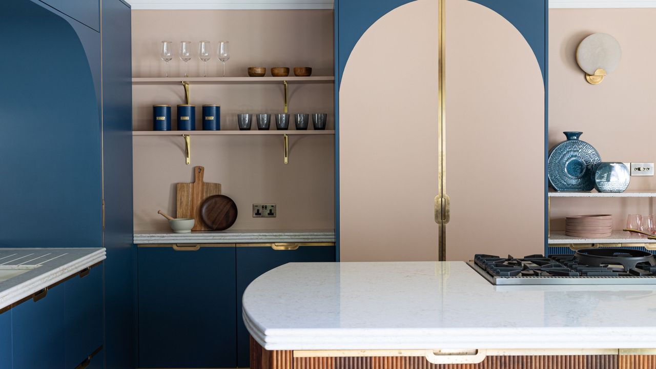 Two-tone kitchen ideas are so chic. Here is a kitchen with dark blue and pale pink walls, wall shelves with blue glassware on, and a white kitchen island with wooden panels