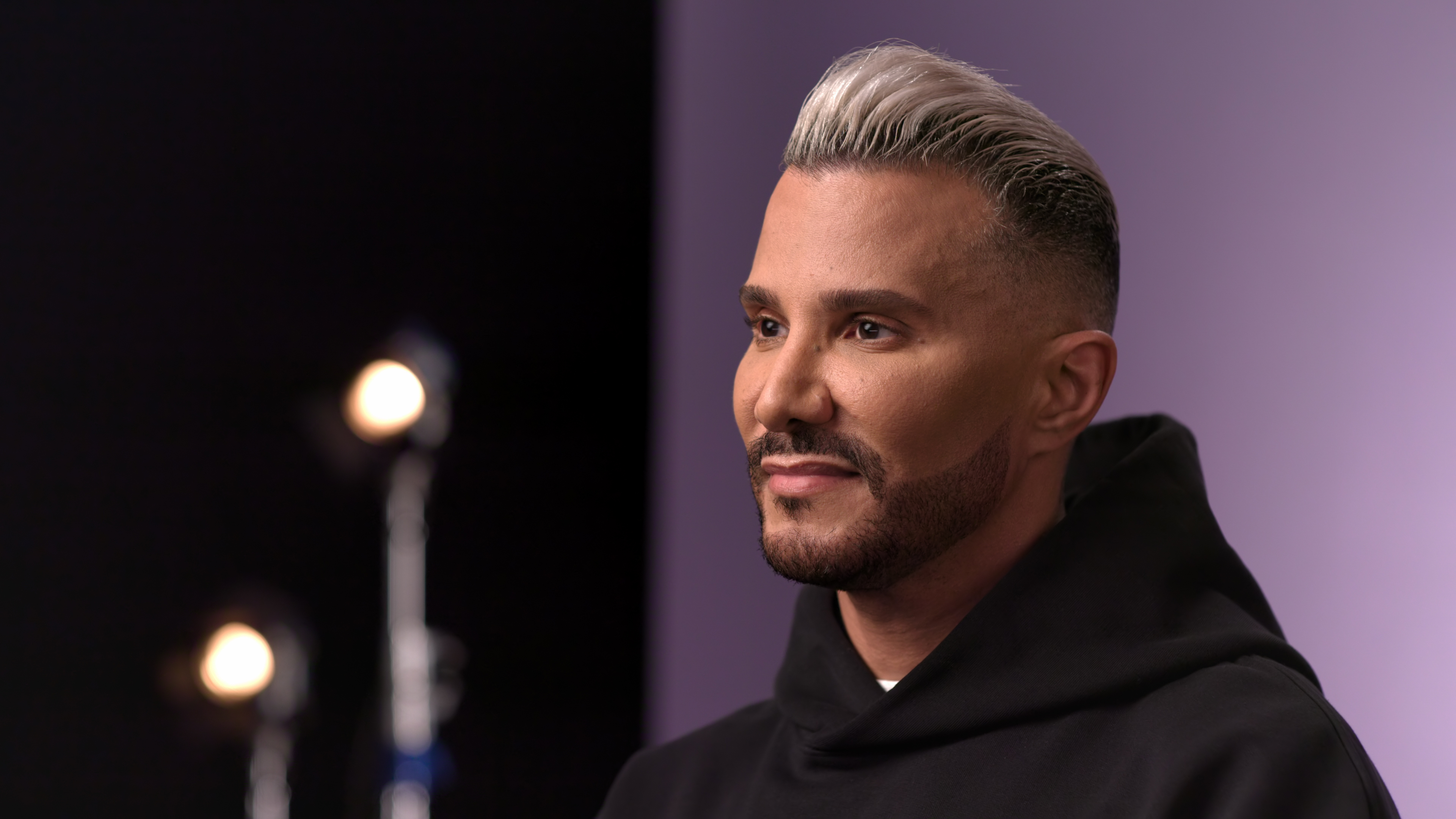 Creative director and TV personality Jay Manuel wears a black hoodie and sits in front of a purple backdrop on a filming set, in the Netflix docuseries 'Reality Check: Inside America's Next Top Model.'