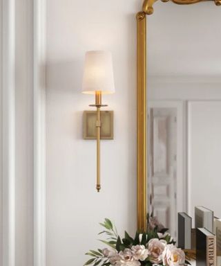 A gold wall sconce on a white wall next to a gold mirror, with flowers and books underneath it