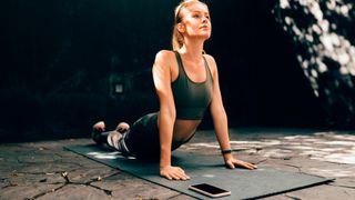 Woman holding upward-facing dog yoga move with smartphone on mat in front of her. She is wearing Adidas UltraBoost running shoes 